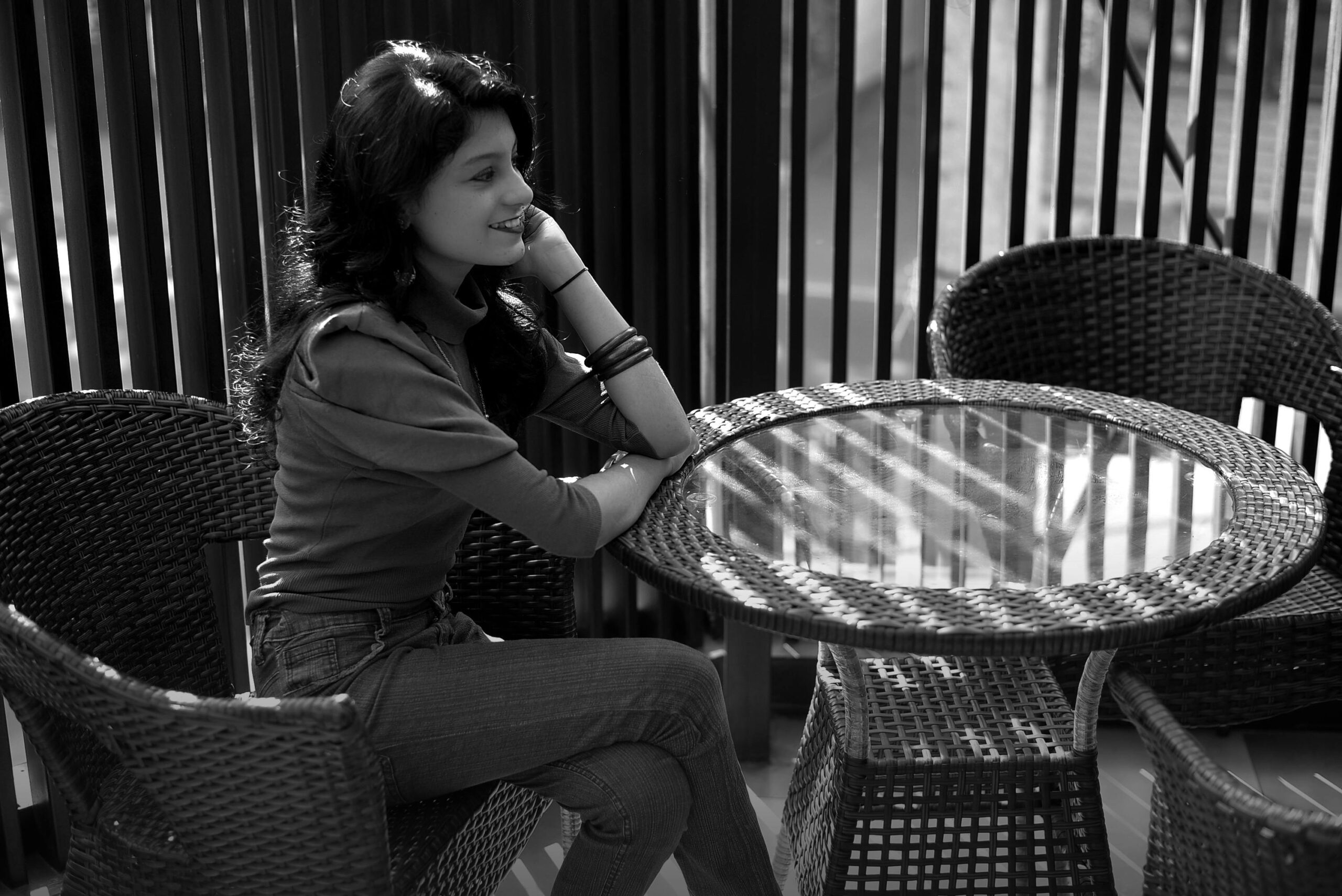Black and white photo of a young woman sitting at an outdoor cafe table, smiling and relaxed.
