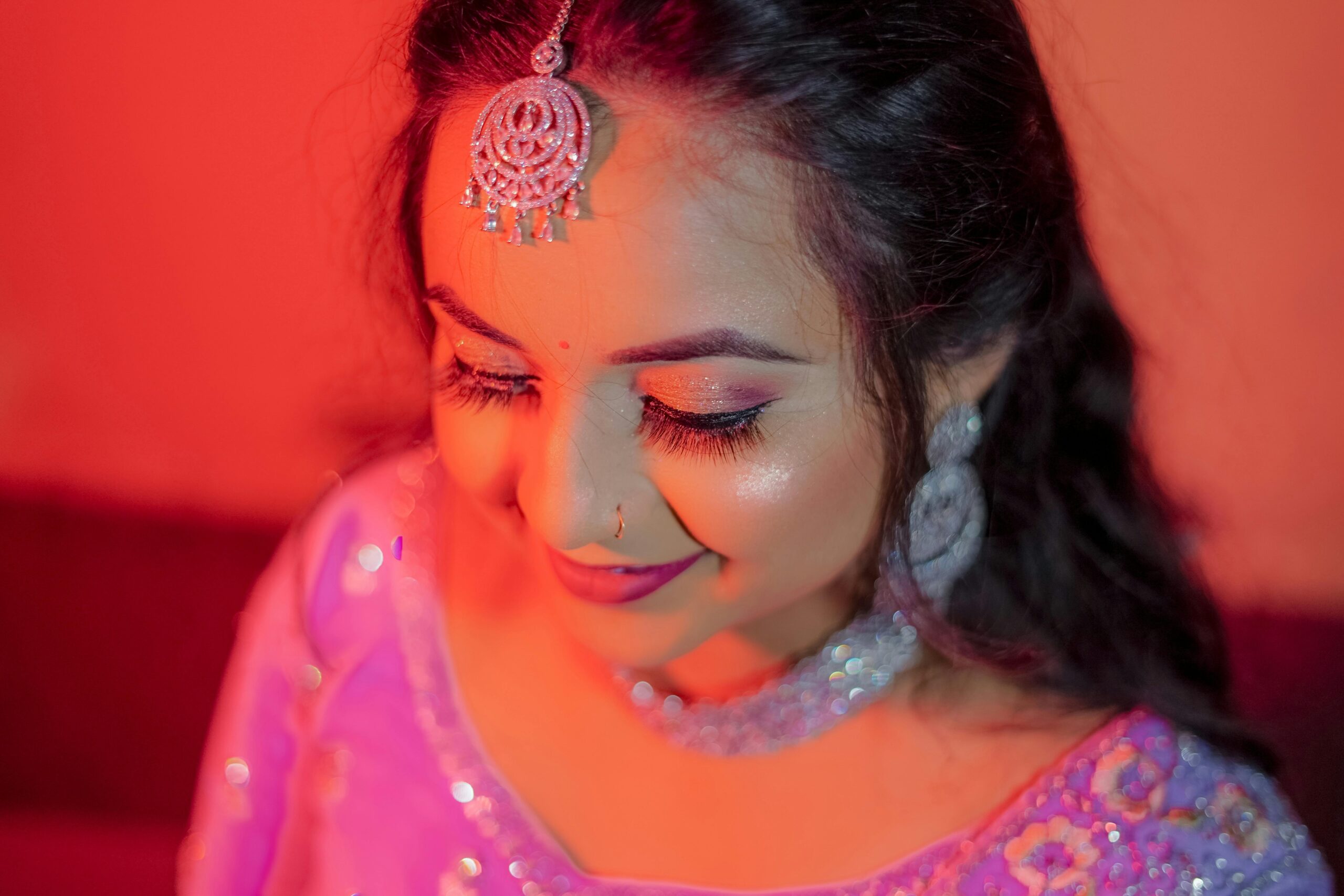 Beautiful woman in traditional Indian attire and jewelry, smiling softly under warm red lighting.